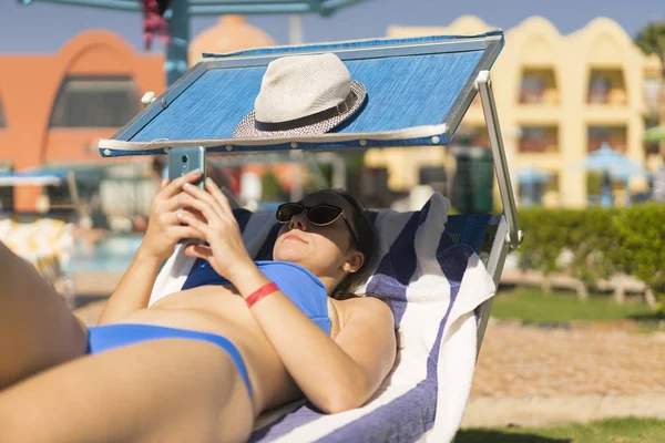 Sommer-Lifestyle-Porträt einer jungen atemberaubenden Frau, die auf der Sonnenliege am Strand der tropischen Insel liegt und ihr Smartphone benutzt. junge weiße Frau auf blauer Sonnenliege liegend — Stockfoto
