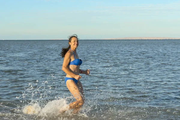 セクシーな若いブルネットの女性または捨てられた熱帯のビーチと青い空でサーフィンを介して実行されているビキニを着ている少女。海沿い走っている若い女性。コピー スペース — ストック写真