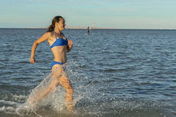 セクシーな若いブルネットの女性または捨てられた熱帯のビーチと青い空でサーフィンを介して実行されているビキニを着ている少女 海沿い走っている若い女性 — ストック写真