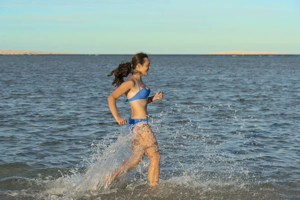 En sexig ung brunett kvinna eller flicka iklädd en bikini som löper genom att surfa på en öde tropisk strand med en blå himmel. Ung kvinna kör vid havet — Stockfoto