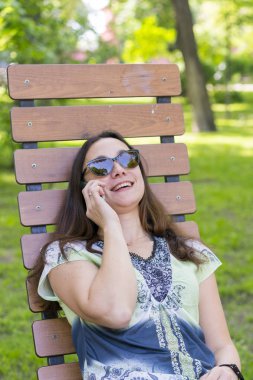 Genç kadın parktaki bankta dinleniyor. Güzel kadın bir park bankta rahatlatıcı ve bir akıllı telefon kullanarak. Tonda