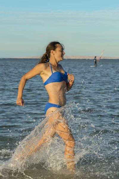 Uma jovem morena sexy usando um biquíni correndo pelo surf em uma praia tropical deserta com um céu azul. Uma jovem a correr à beira-mar. foto vertical — Fotografia de Stock