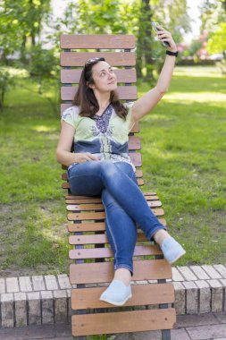 Genç kadın parkta selfie yapıyor. Uzun sarı saçlı seksi genç kadın parkta yaz gününde selfie yapıyor. dikey fotoğraf