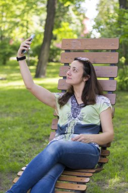 Genç kadın parkta selfie yapıyor. Uzun sarı saçlı seksi genç kadın parkta yaz gününde selfie yapıyor. Neşeli kız parkta yaz gününde selfie yapma. dikey fotoğraf.