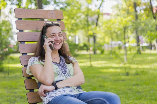 Молодая женщина разговаривает по смартфону и смеется в парке на скамейке Красивая женщина отдыхает на скамейке в парке и пользуется смартфоном. тонированные — стоковое фото