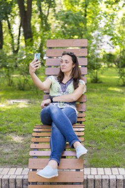 Genç kadın parkta selfie yapıyor. Uzun sarı saçlı seksi genç kadın parkta yaz gününde selfie yapıyor. dikey fotoğraf
