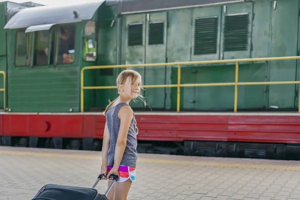Dziewczyna nastolatek na stacji kolejowej w pobliżu lokomotywy. Dziewczyna ciągnie dużą walizkę na platformie — Zdjęcie stockowe