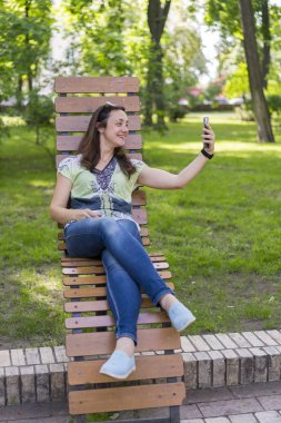 Genç kadın parkta selfie yapıyor. Uzun sarı saçlı seksi genç kadın parkta yaz gününde selfie yapıyor. dikey fotoğraf