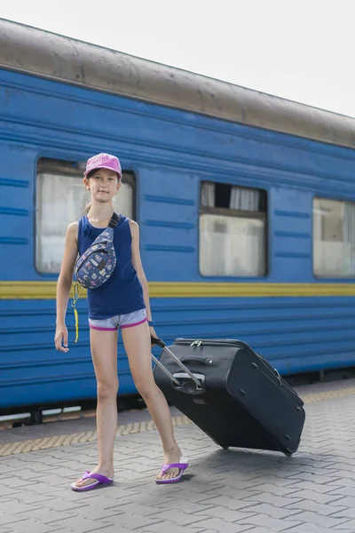 Mała słodka dziewczyna z dużą walizką na opuszczonej platformie kolejowej. Dziewczyna ciągnąc dużą walizkę na platformie. Zdjęcie pionowe — Zdjęcie stockowe