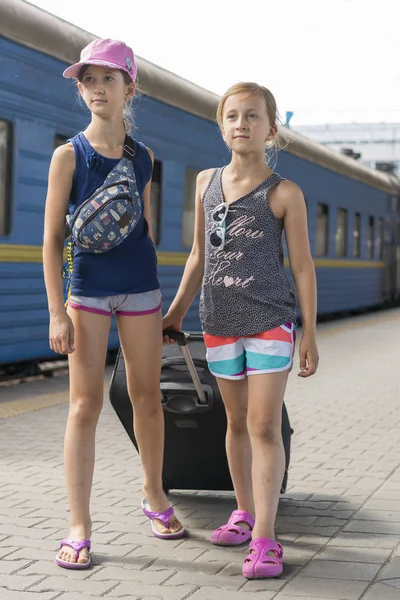 Dwie nastoletnie dziewczyny na dworcu kolejowym z walizka. Dwie siostry ciągną duży i ciężki czarny Walizka na tle pociągu. Zdjęcie pionowe — Zdjęcie stockowe