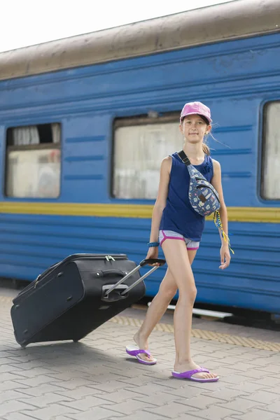 Mała słodka dziewczyna z dużą walizką na opuszczonej platformie kolejowej. Dziewczyna ciągnąc dużą walizkę na platformie. Zdjęcie pionowe — Zdjęcie stockowe