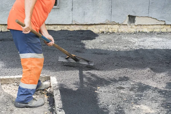 Hombre trabajador pone asfalto carretera reparación carretera pavimentación luz de rayos de sol amarillo. Un hombre en overol está poniendo asfalto con una pala — Foto de Stock