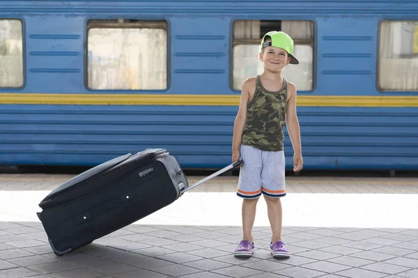 Счастливый маленький мальчик с большим черным чемоданом на фоне синего поезда. Концепция приключений и путешествия — стоковое фото