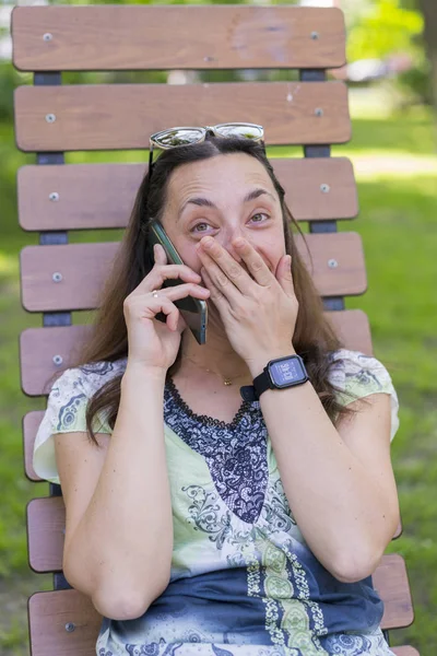 Молодая женщина разговаривает на смартфоне и смеется в парке на скамейке. Красивая женщина отдыхает на скамейке в парке и пользуется смартфоном. — стоковое фото