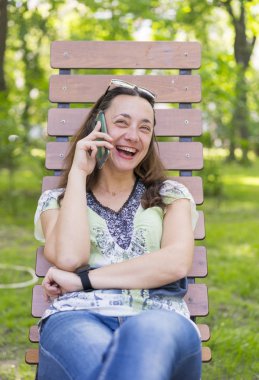 Akıllı telefonda konuşan ve bankta parkta gülen genç kadın.