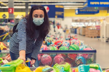 Süpermarkette koruyucu maskeli bir kadın çocuk oyuncakları seçiyor. ses tonlu.