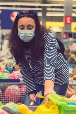 Süpermarkette koruyucu maskeli bir kadın çocuk oyuncakları seçiyor. Ton gibi. Dikey fotoğraf. ses tonlu.