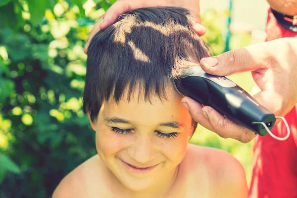 hand holds an electric hair clipper and cuts the boy\'s long hair, barbershop at home, parent cuts hair while hairdressers are closed, stay home concept. Smiling boy. toned.