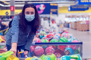 Süpermarkette koruyucu maskeli bir kadın çocuk oyuncakları seçiyor. ses tonlu.