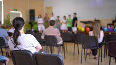 Anaokulundaki küçük çocuklar ailelerinin önünde sahneye çıkıyorlar. Birçok ebeveyn koridorda çocukların performansını izliyor. Sahnedeki çocuklar ailelerinin önünde gösteri yapıyorlar. Bulanık. ses tonlu