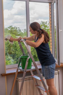Odada pencereyi paçavra ve cam temizleyicisiyle temizleyen genç kadının arka görüntüsü. Uzun saçlı tatlı kız bardağı deterjanla yıkıyor. seçili odak.