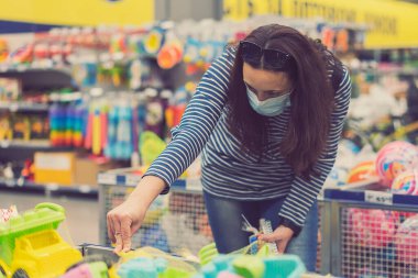Süpermarkette koruyucu maskeli bir kadın çocuk oyuncakları seçiyor. ses tonlu.