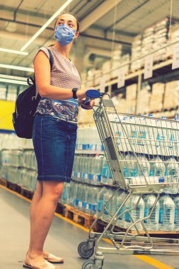 Telaşlı kadın, süpermarkette ya da mağazada alışveriş yaparken Coronavirus 'a karşı tıbbi maske takar. Sağlık, güvenlik ve salgın konsepti. Alışveriş merkezinde alışveriş kolik bir kız. Dikey fotoğraf. ses tonlu.