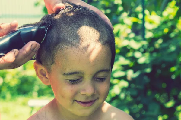 hand holds an electric hair clipper and cuts the boy\'s long hair, barbershop at home, parent cuts hair while hairdressers are closed, stay home concept. toned.