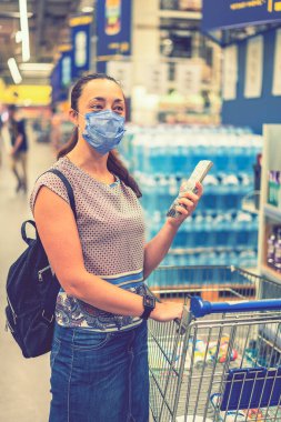 Telaşlı kadın, süpermarkette ya da mağazada alışveriş yaparken Coronavirus 'a karşı tıbbi maske takar. Sağlık, güvenlik ve salgın konsepti. Alışveriş merkezinde alışveriş kolik bir kız. Dikey fotoğraf. ses tonlu.