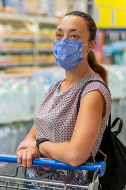Telaşlı kadın, süpermarkette ya da mağazada alışveriş yaparken Coronavirus 'a karşı tıbbi maske takar. Sağlık, güvenlik ve salgın konsepti. Alışveriş merkezinde alışveriş kolik bir kız. Dikey fotoğraf. Ton gibi. Kapat..
