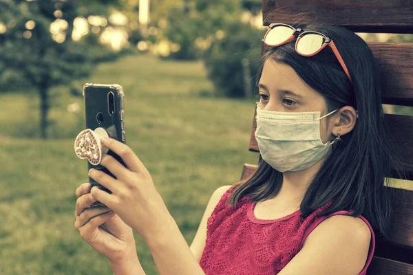 Akıllı telefon tutan koruyucu maskeli bir kız. Green Park 'taki bankta oturan genç bir kız. Cep telefonuyla. Koronavirüs kısıtlamalarının gevşetilmesi ve kendini izole etmenin ilk aşaması. ses tonlu — Stok fotoğraf