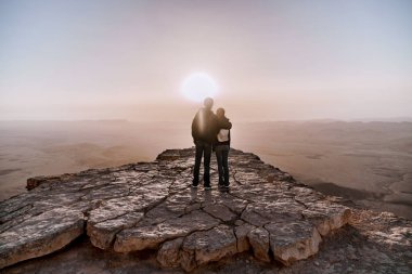 Alone young couple of man and women with backpack in israel negev desert admires the view of sunrise. Young pair stands on the edge of the cliff of makhtesh ramon park. clipart