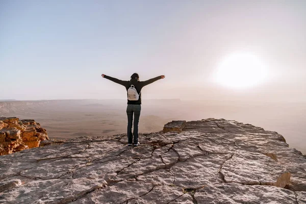 Solo joven con mochila en el desierto de Israel Néguev admira la vista del amanecer. Joven mujer se encuentra en el borde del acantilado del parque Makhtesh Ramon . — Foto de Stock