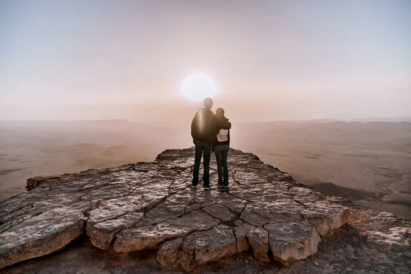 男とバックパックで女性の一人で若いカップル イスラエルのネゲヴ砂漠日の出の景色を賞賛します。Makhtesh の崖の端に立っている若いペア ラモン公園. — ストック写真