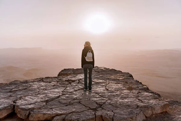 Solo joven con mochila en el desierto de Israel Néguev admira la vista del amanecer. Joven mujer se encuentra en el borde del acantilado del parque Makhtesh Ramon . — Foto de Stock