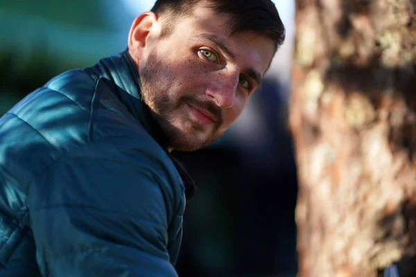 Güneşin ışıklarında bir ağacın bulanık arka plan üzerinde mavi ceket kısa kahverengi saçlı ve yeşil gözlü çilleri ile gülümseyen sakallı adam Portresi. Erkek kameraya bakıyor..