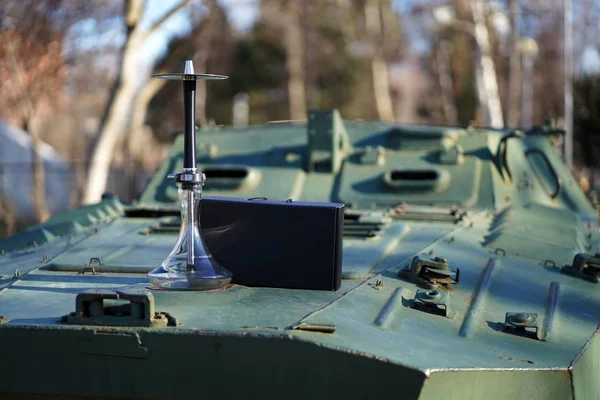 Ormanın arka planında güneş ışığı bir askeri koyu yeşil zırhlı personel taşıyıcı üzerinde bir kutu ile temiz bir şeffaf kase ile lüks siyah mat nargile. Askeri dinlenme kavramı.