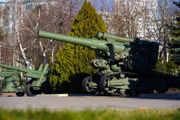 B-4 - a 203-mm Soviet green with rusty stains howitzer of the 1931 model among the trees next to the old Russian military equipment in the rays of the sun against the backdrop of modern buildings. — Stock Photo, Image