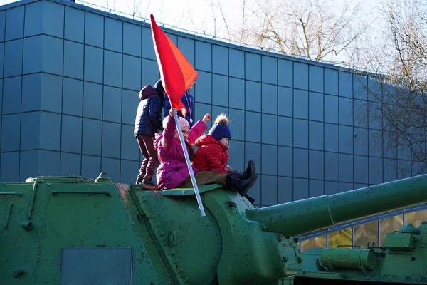 Renkli ceket ve şapkalı küçük çocuklar eski yeşil tankın üzerinde oturup koyu mavi binanın arka planında keyfini çıkarırlar. Pembe ceketli kız SSCB'nin kırmızı bayrağını dalgalandırıyor ve kutlama yapıyor..