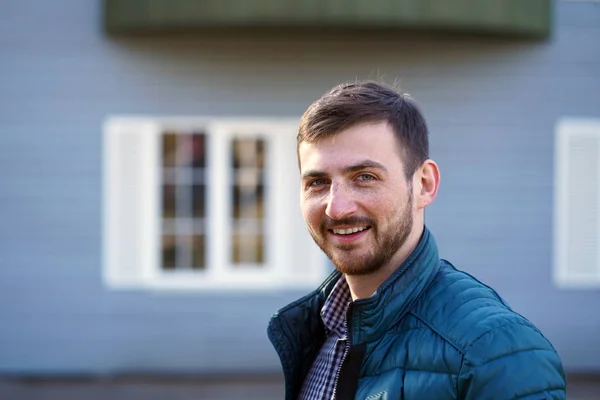 Portrait of a laughing bearded man with freckles with short brown hair and green eyes in a blue jacket and plaid shirt on the background of a blue house with white windows in the sun. Stock Image