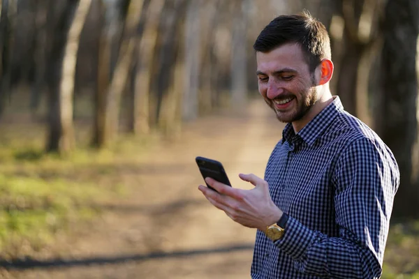 Kısa kahverengi saçlı çiller ve altın saat ile ekose gömlek yeşil gözlü çekici sakallı kafkas adam telefonda bir mesaj okur ve güneş ışınları sonbahar ormanda gülüyor.