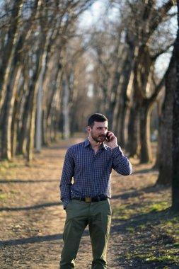 Ekose düğmesiz gömlek kısa saçlı çiller ile İş sakallı adam, güneşte ağaçların arasında sonbahar ormanında cebinde eliyle telefonda konuşurken zeytin pantolon.