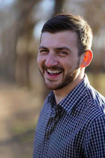 Bulanık bir orman arka plan karşı güneşin altında ekose unbuttoned gömlek kısa kahverengi saçlı ve yeşil gözlü çiller ile gülen sakallı kafkas bir adam portresi. Erkeğin kırmızı kulağı var..