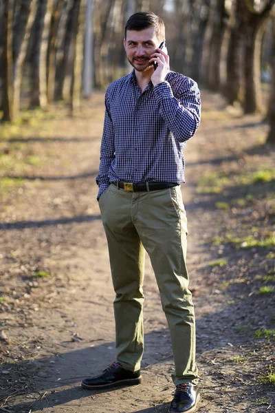 Ekose gömlek, zeytin pantolon ve siyah ayakkabı kısa kahverengi saçlı çiller ile Iş sakallı kafkas adam ağaçların arasında sonbahar ormanında cebinde eliyle telefonda konuşuyor.