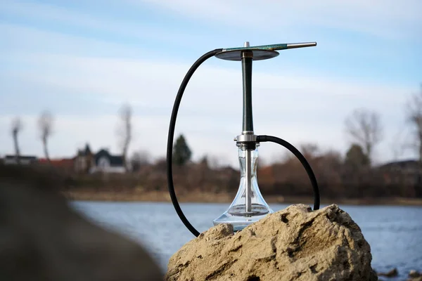 Koyu bir hortum ile berrak su ile mavi ahşap nargile bir taş üzerinde nehir yakınında doğada duruyor. Seyahat ve nargile sigara kavramı. Bulutlu bir gökyüzü ve evlerin siluetleri ile bulanık arka plan.