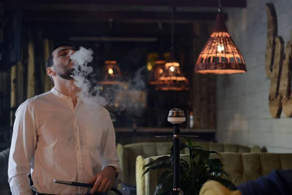 Zevkle siyah pantolon beyaz gömlek çiller ile Sakallı adam bir mat nargile sigara sahiptir ve lounge barda ağzından duman darbe. Restoranın içi. Dinlenme kavramı.