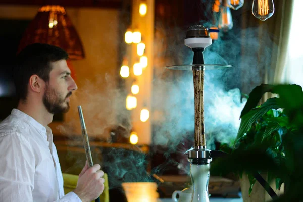Zevkle beyaz gömlekli sakallı bir adam bir lounge barda bej ahşap nargile içiyor. Erkek ağızlığı tutar ve duman üfler. Restoranın içi. Dinlenme kavramı.