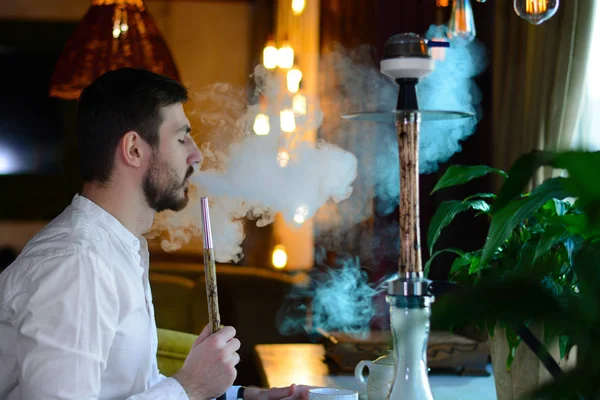 A bearded man in white shirt drinks tea and smokes with pleasure a beige wooden hookah with lightning patterns in a lounge bar. Male make a cloud of smoke from mouth. Concept of rest. Royalty Free Stock Photos