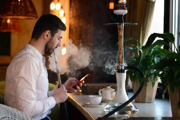 Beyaz gömlekli sakallı genç bir adam yıldırım desenli bej ahşap nargile içiyor ve telefona bakıyor, salonda bir mesaj yazdırıyor. Restoranın içi. Dinlenme kavramı.