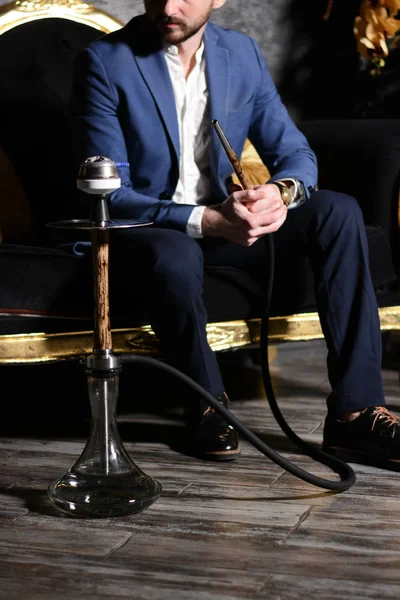 Beyaz gömlekli, mavi ceketli ve koyu renk pantolonlu sakallı bir adam kanepeye oturur ve evde elektrik akımından kaynaklanan ışık hızında desenleri olan bej ahşap nargile içer. Dinlenme kavramı.